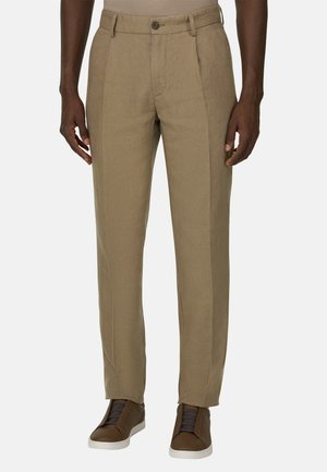 Homme portant un pantalon beige sur mesure et des baskets marron à semelles blanches, debout devant un fond uni.