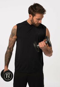 Maglietta atletica nera senza maniche, che tiene dei manubri neri, con dettagli dei tatuaggi visibili sulle braccia, su uno sfondo bianco semplice.