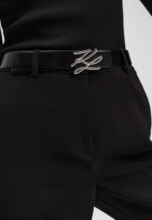 Cintura in pelle nera con una fibbia in metallo argentato dal design astratto, indossata sopra pantaloni neri con una trama liscia.