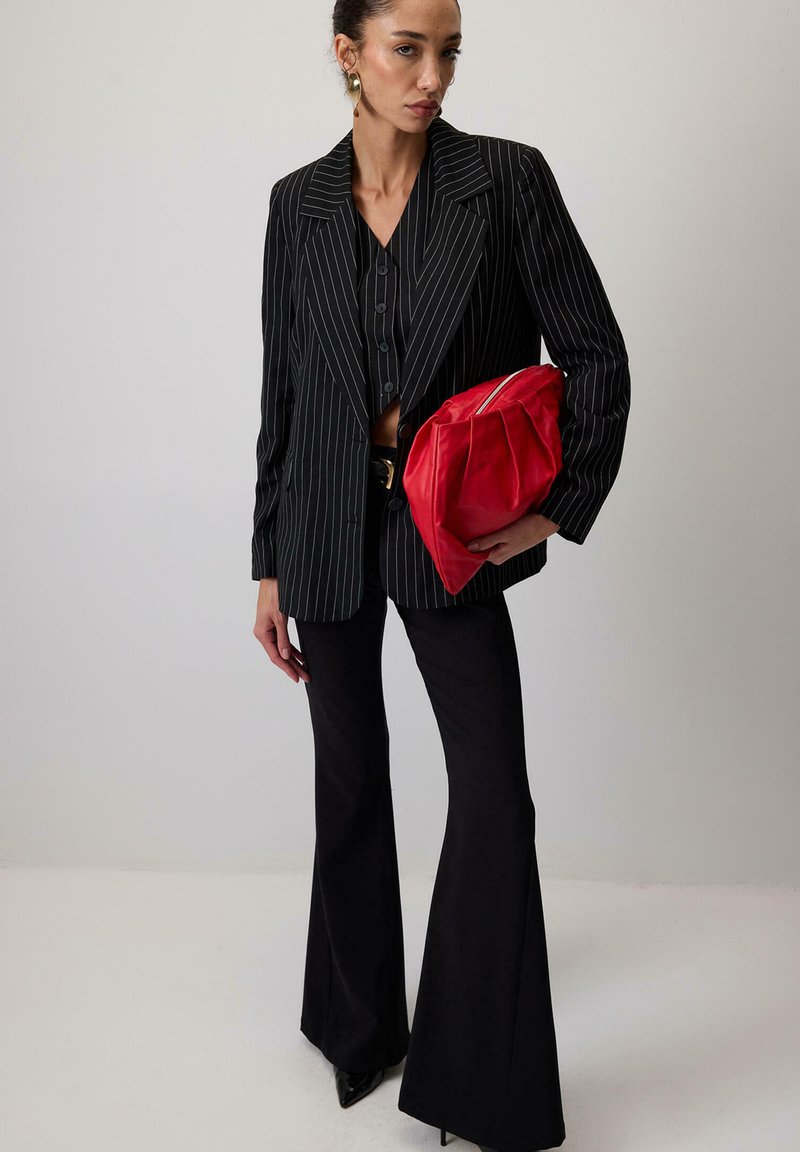 Sacou negru cu dungi fine peste o cămașă neagră scurtă cu butoane, asociat cu pantaloni negri evazați și o poșetă roșie cu un design plisat.
