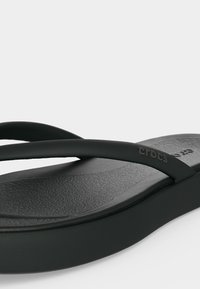 Κοντινή λήψη ενός μαύρου Crocs σαγιονάρας με υφή στην πάτο και λεπτά λουράκια, με το λογότυπο Crocs στο πλευρικό λουράκι.