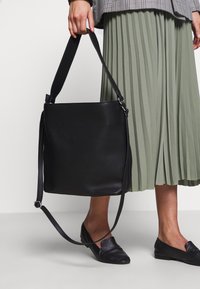 Persona che indossa mocassini neri e una gonna a pieghe verde, mentre tiene una grande borsa in pelle nera con tracolle.