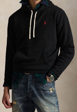 Personne portant un sweat à capuche noir avec des cordons blancs, une chemise à carreaux, un jean bleu usé et une montre, debout avec une main dans une poche.