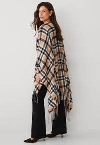 Plaid poncho in beige, black, and red, features fringed edges and a loose design. Paired with black flared pants and heels.