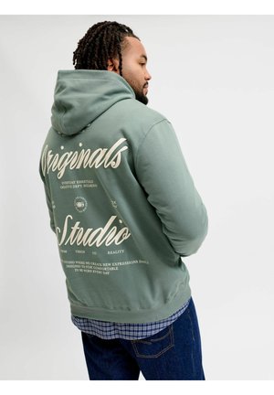 Man with braided hair wearing green hoodie with "Originals Studio" text on back, blue jeans, and checkered shirt underneath, facing sideways.