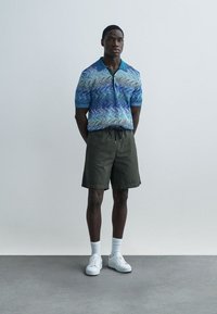 Blaues Polohemd mit Wellenmuster, kombiniert mit olivgrünen Shorts. Das Modell trägt weiße Sneakers und Kniestrümpfe.