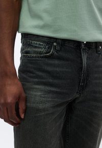 Schwarze Jeans aus Denim mit ausgewaschenem Look, Fünf-Taschen-Design und Metallknopfverschluss. Kombiniert mit einem hellgrünen Hemd, das eine strukturierte Stoffqualität zeigt.