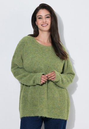 Kvinde med langt mørkt hår iført en blød grøn sweater og blå bukser, smilende med hænder løst sammenfoldet foran en hvid baggrund.