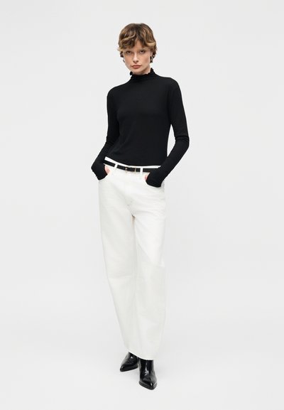 Άτομο που φοράει μαύρο μπλουζάκι με μακριά μανίκια, λευκό παντελόνι φαρδύ στο κάτω μέρος με λευκή ζώνη, μαύρες μυτερές μπότες, στέκεται με τα χέρια στις τσέπες.