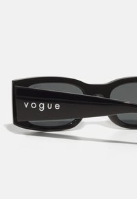 Ochelari de soare negri cu un design geometric masiv, lentile întunecate și numele brandului "vogue" în alb pe brațul lateral. Finisaj lucios.