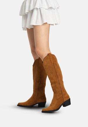 Benen met kniehoge bruine suède cowboylaarzen met lage blokhakken, gecombineerd met een korte witte rok met lagen.