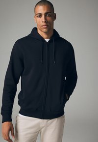 Haut à capuche noir zippé en matériau doux, avec un cordon de serrage, des poignets côtelés et une coupe décontractée. Porté avec un pantalon de couleur claire.