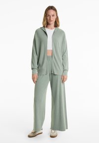 Ljusgrön stickad zip-up hoodie med ribbat mönster, kombinerad med vida stickade byxor i samma material. Vit cropped topp och sneakers.