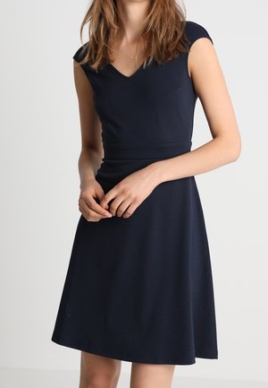 Femme portant une robe bleu marine sans manches, longueur au genou, avec un col en V, debout devant un fond clair uni, les mains légèrement jointes.