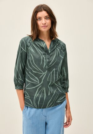 Blusa verde oscura con escote en V y mangas de tres cuartos, con un patrón de hojas en un verde más claro. Con tapeta delantera con botones.