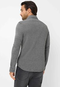 Langärmliges graues Hemd mit strukturierter Oberfläche, geknöpften Manschetten und einem entspannten Kragen. Getragen mit dunkelgrauen Jeans, von hinten gezeigt.