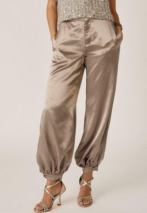 Pantalones de satén sueltos con puños elásticos, combinados con sandalias de tacón alto con tiras y un top de lentejuelas, con las manos en los bolsillos.