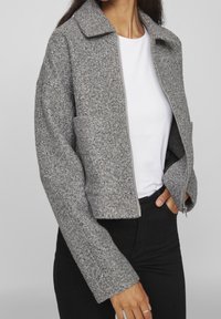 Szara, teksturowana kurtka zapinana na zamek z kołnierzem i dwoma przednimi kieszeniami, w zestawie z białą koszulą i czarnymi spodniami.