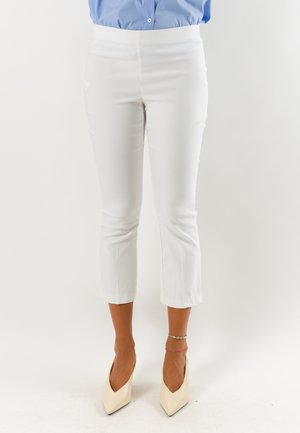 Parte inferiore del corpo di una persona che indossa pantaloni bianchi corti, tacchi beige appuntiti e cavigliere su uno sfondo chiaro e uniforme.