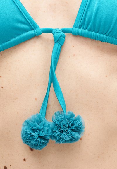 Κοντινό πλάνο στην πλάτη ενός ατόμου που φοράει κορυφή μαγιό μπικίνι σε τιρκουάζ χρώμα, δεμένη με κορδόνια από τα οποία κρέμονται διακοσμητικά πομ-πομ.