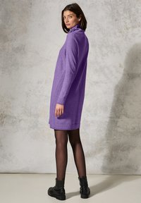 Abito maglione viola con colletto alto, maniche lunghe e dettagli a costine. Indossato con stivaletti neri alla caviglia, su uno sfondo grigio chiaro testurizzato.