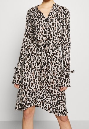 Kvinde iført en beige leopardprint langærmet kjole med bindebælte i taljen og asymmetrisk kant mod en ensfarvet baggrund.