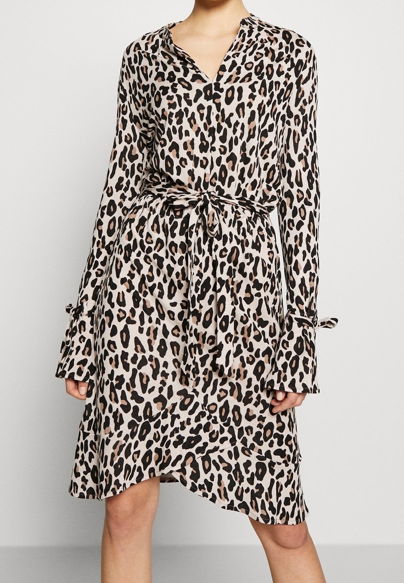 Femme portant une robe beige à imprimé léopard, manches longues, ceinture nouée à la taille et ourlet asymétrique, sur un fond uni.