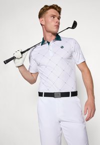 Hvid golf polo med en mørkegrøn krave, med et diamantmønster. Bæres med hvide bukser og et sort bælte, mens der holdes en golfkølle.