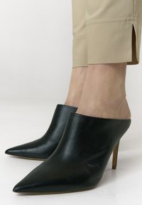 Schwarze Mules aus Leder mit spitz zulaufender Vorderseite und hohem Absatz. Glatte Oberfläche mit kleinen Details. Kombiniert mit beigen, schmal zulaufenden Hosen.