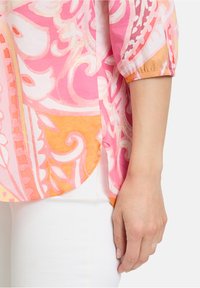 Blouse à motifs rose et orange avec une coupe ample, des manches trois-quarts et un ourlet asymétrique, fabriquée en tissu léger.