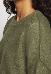 Pull vert olive en matériau doux et pelucheux. Dispose d'un col rond et de manches raglan, mettant en valeur une finition texturée.