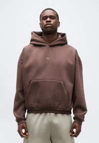 Νεαρός άντρας που φοράει καφέ φουτέ με μικρό λευκό λογότυπο και ανοιχτό μπεζ παντελόνι, όρθιος με τα χέρια χαλαρά δίπλα στα πλευρά.