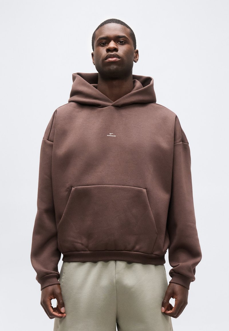 Νεαρός άντρας που φοράει καφέ φουτέ με μικρό λευκό λογότυπο και ανοιχτό μπεζ παντελόνι, όρθιος με τα χέρια χαλαρά δίπλα στα πλευρά.