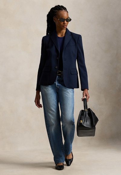 Γυναίκα με πλεξούδες, φοράει σκούρα γυαλιά ηλίου, μπλε σακάκι, μπλε τζιν, μαύρα παπούτσια και κρατά μια μεγάλη μαύρη τσάντα σε ουδέτερο φόντο.