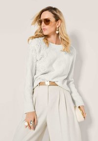 Frau in einem weißen strukturierten Pullover, beigen hochgeschnittenen plissierten Hosen, mit einem braunen Gürtel, übergroßen braunen Sonnenbrillen, Perlenohrringen und einer weißen Clutch.
