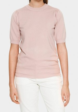 Kvinde iført en lys pink kortærmet striktop med puffede skuldre og hvide bukser, stående mod en ensfarvet baggrund.