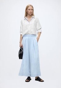 Femme aux longs cheveux blonds portant une chemise blanche ample, un pantalon large bleu clair, des sandales noires et un sac à main noir, debout sur fond blanc.
