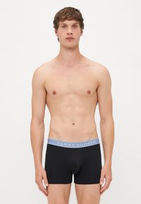 Homem jovem em pé contra um fundo branco, vestindo cuecas boxer pretas da Tommy Hilfiger com cós azul claro.