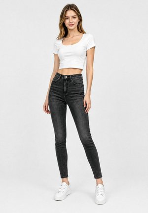 Ung kvinde iført en hvid cropped top, sorte højtaljede skinny jeans og hvide sneakers, stående mod en ensfarvet baggrund.