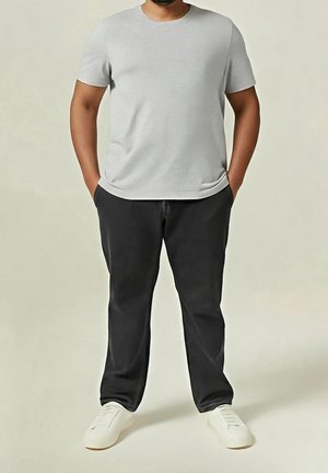 Homme portant un t-shirt gris clair uni à manches courtes, un pantalon noir et des baskets blanches, debout les mains dans les poches.