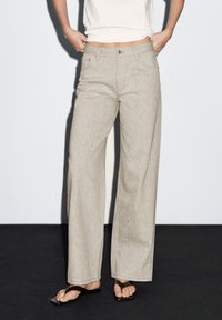 Beige weit geschnitte Hose mit Knopf- und Reißverschluss, zwei vorderen Taschen und einer subtilen melierten Textur.