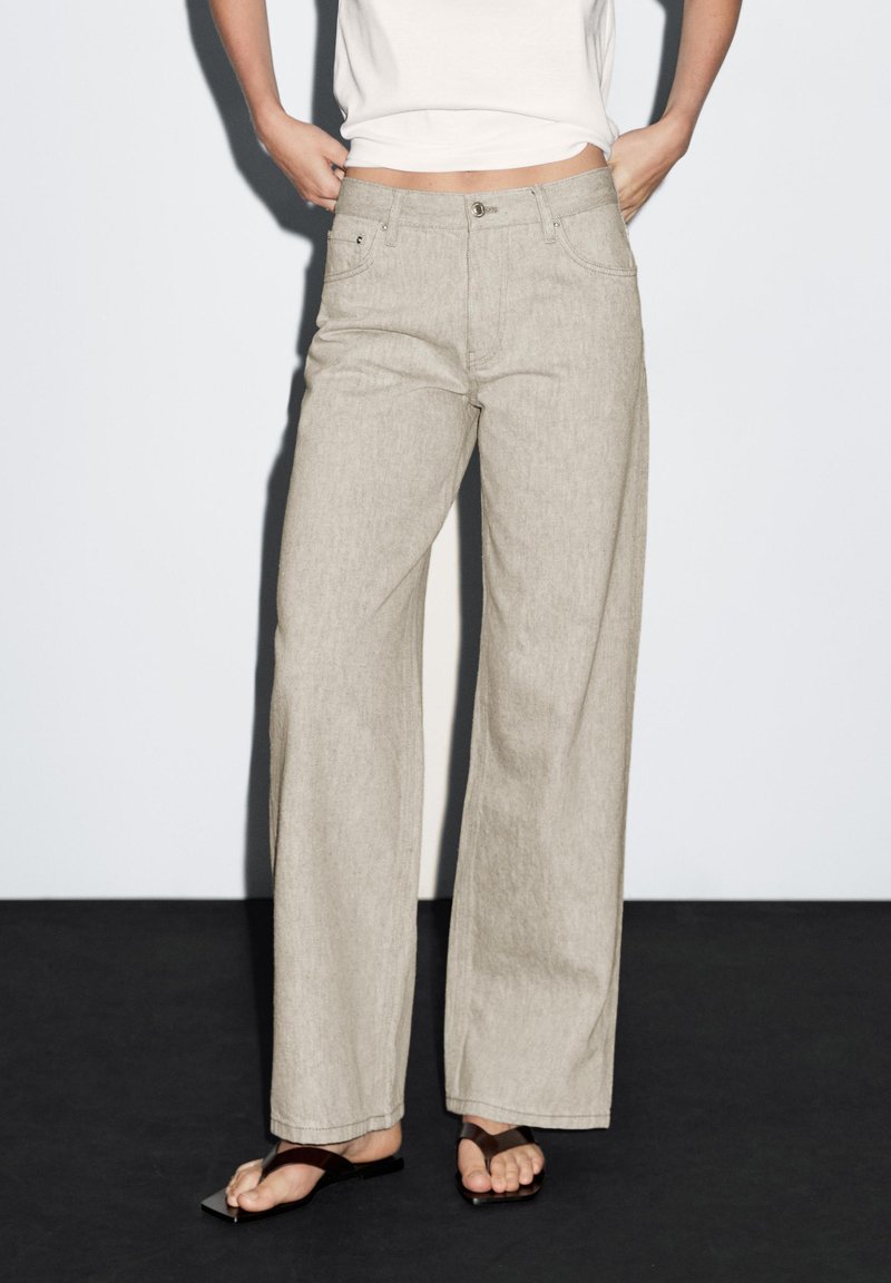 Beige weit geschnitte Hose mit Knopf- und Reißverschluss, zwei vorderen Taschen und einer subtilen melierten Textur.