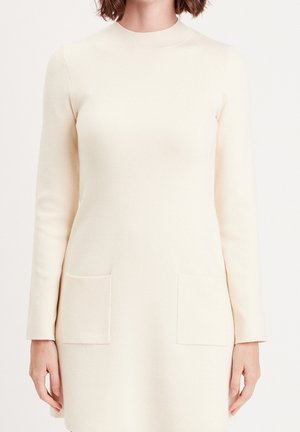 Robe en maille à manches longues couleur crème avec col rond et deux poches plaquées devant, portée par une personne aux cheveux mi-longs.