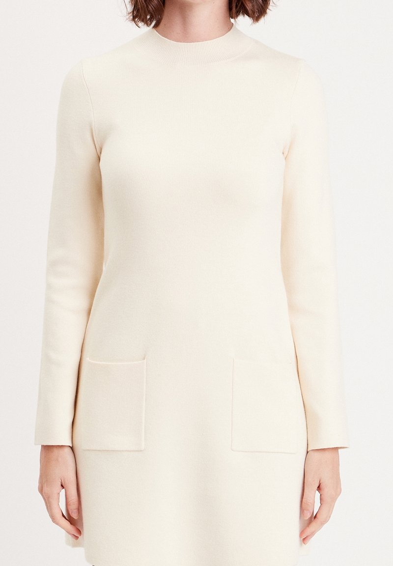 Robe en maille à manches longues couleur crème avec col rond et deux poches plaquées devant, portée par une personne aux cheveux mi-longs.