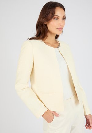 Mujer de cabello castaño lleva un blazer crema sin cuello sobre una camiseta blanca, con una mano en el bolsillo del blazer, mirando ligeramente hacia la derecha.