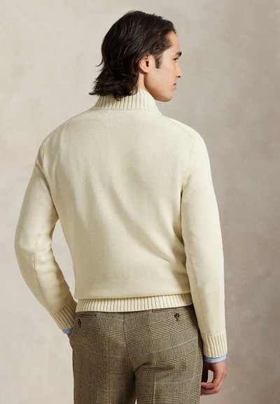 Keskmise pikkusega tumedate juustega mees, kes kannab koorevärvi kõrge kaelusega kampsunit ja pruune ruudulisi pükse, seisab beige tausta ees seljaga vaatajapoolt.