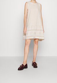Ärmelloses beiges Kleid mit Hahnentrittmuster, gerafftem Bund und geschichtetem Saum. Die Schuhe sind dunkelburgunderfarbene Slipper mit einem goldenen Schnallen-Detail.