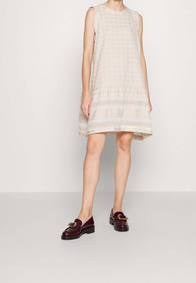 Ärmelloses beiges Kleid mit Hahnentrittmuster, gerafftem Bund und geschichtetem Saum. Die Schuhe sind dunkelburgunderfarbene Slipper mit einem goldenen Schnallen-Detail.