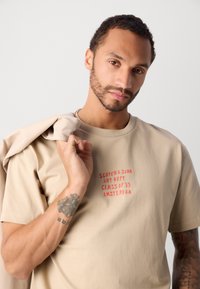 Beige pamut póló piros "SCOTCH & SODA ART DEPT. CLASS OF '85 AMSTERDAM" felirattal, kerek nyakkivágással és laza szabással.