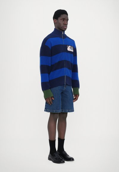 Knitted zip-up sweater with wide blue and navy stripes, features a hand design on chest pocket. Paired with denim shorts and black loafers.